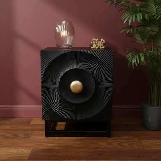 Black decorative cabinet with gold ball on a wooden floor against a pink wall.