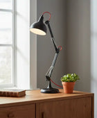 Black adjustable desk lamp on a wooden surface with a potted plant and book in a room with a window.