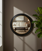 Round wall mirror with a black ribbed frame and gold inner trim, mounted on a taupe wall in a styled interior. The mirror reflects a living room with a cream sofa and coffee table, and a green potted plant stands to the right.