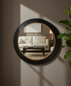 A round wall mirror with a thick black ribbed frame and a thin gold inner trim, mounted on a warm taupe wall. The mirror reflects a cozy living room scene with a cream sofa and a dark coffee table.