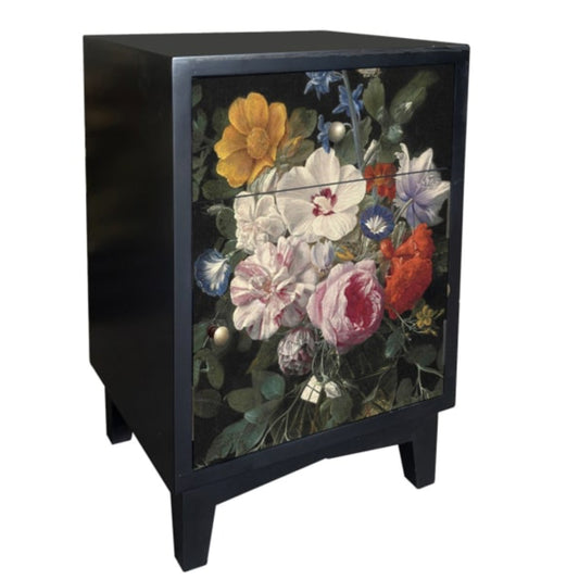 A black wooden side table or cabinet with two drawers, featuring a detailed floral print on the front doors. The flowers include yellow, white, pink, red, and blue blossoms with green leaves, set against a dark background.