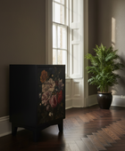 A dark wooden cabinet with a vintage-style floral print on the front, placed in a room with dark wood floors, a tall potted fern, and a tall window.