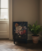 A dark wooden cabinet with a vibrant, colorful floral print on the front, styled in a room corner next to a potted fern on a herringbone wood floor.