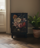A dark cabinet or side table with a large floral artwork panel on the front, styled in a room next to a potted fern on a wood floor.