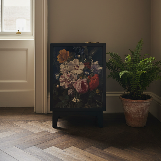 A dark cabinet or side table with a large floral artwork panel on the front, styled in a room next to a potted fern on a wood floor.