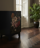 A black two-drawer cabinet with a vintage-style floral print on the front, set in a room with dark wood herringbone floors and a large potted fern nearby.