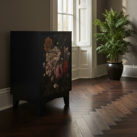 A black two-drawer cabinet with a vintage-style floral print on the front, set in a room with dark wood herringbone floors and a large potted fern nearby.