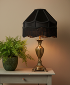 A classic table lamp with a black fringed fabric shade and an ornate gold-toned metal base, styled on a light gray wooden table next to a green potted fern against a warm beige wall.