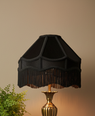 A black fabric lampshade with decorative trim and fringe, mounted on a gold-toned lamp base against a warm beige wall, with a small green plant at the lower left.