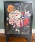 A black wooden nightstand with two doors featuring a large, colorful floral print on the front, set against a muted green wall and wooden floor.