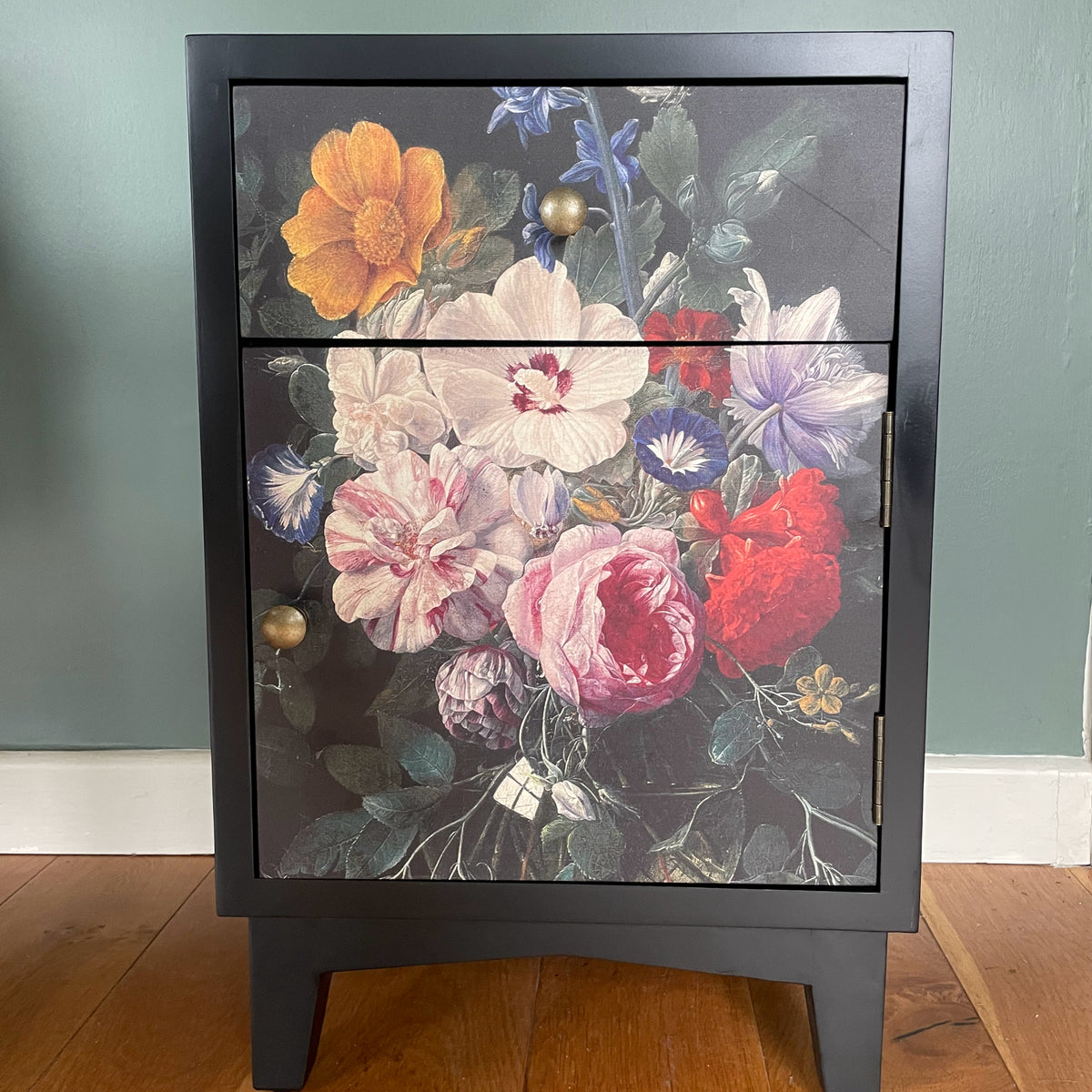 A black wooden nightstand with two doors featuring a large, colorful floral print on the front, set against a muted green wall and wooden floor.