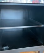 A black wooden sideboard or cabinet with two shelves, shown from the front interior angle. The cabinet has a silver metal hinge at the bottom left and is photographed in a home setting with a floral-patterned curtain and wooden floor visible in the background.