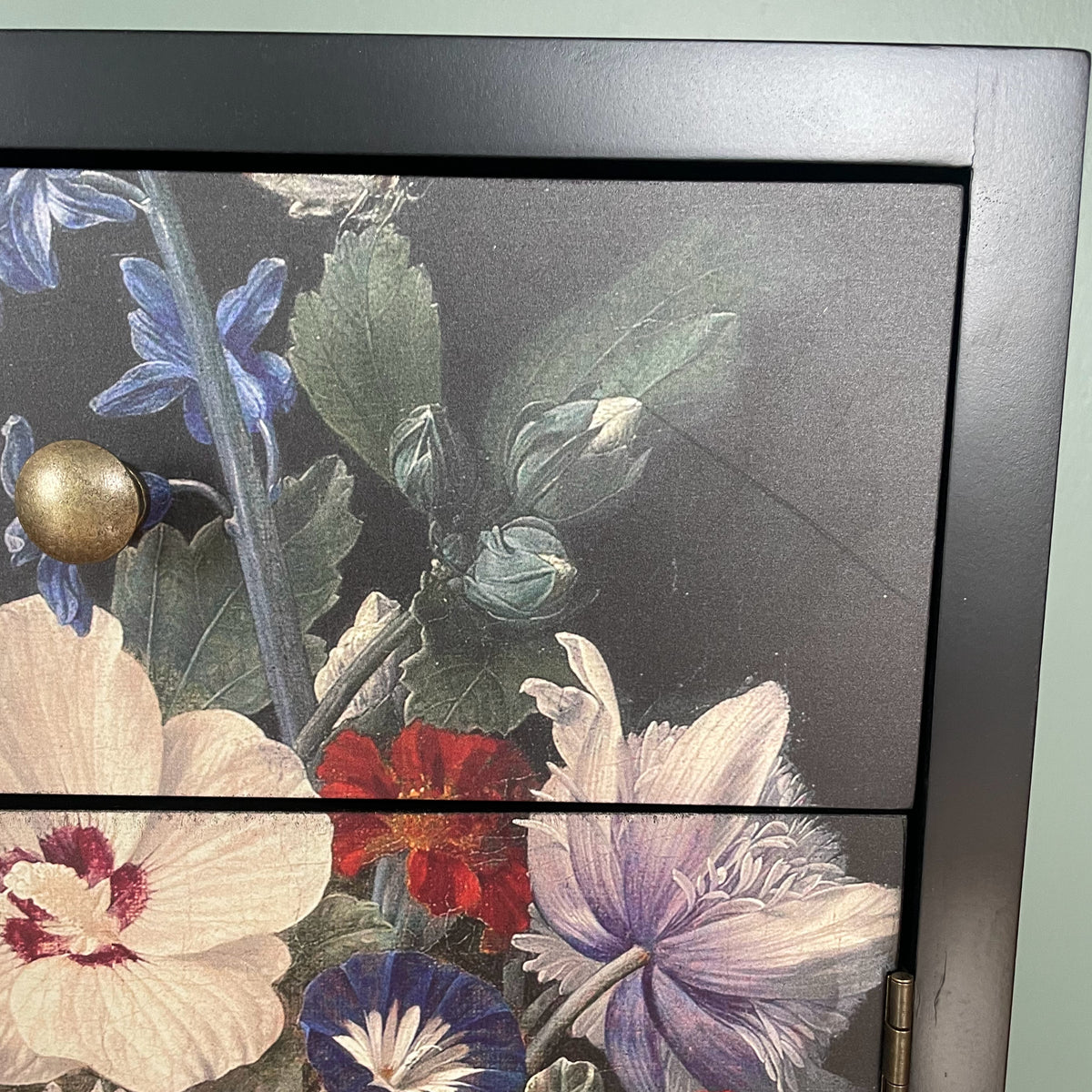Close-up of a dark wood cabinet or drawer front featuring a floral print with blue, red, cream, and purple flowers on a dark background, with a round brass knob visible.