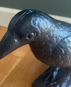 A dark cast iron raven figurine on a matching base, photographed on a wooden floor against a dark green wall.