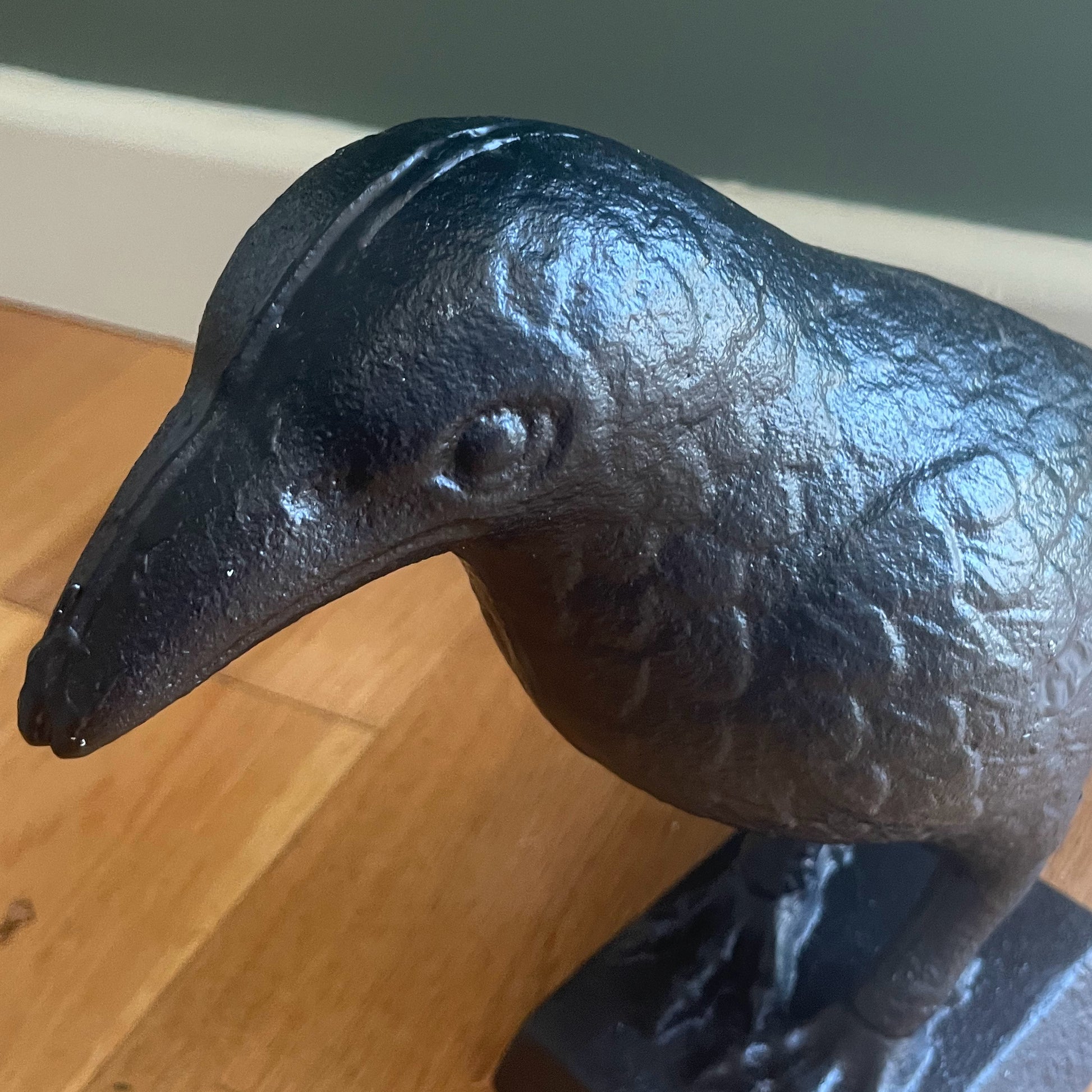 A dark cast iron raven figurine on a matching base, photographed on a wooden floor against a dark green wall.