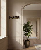 Decorative sign reading 'First Class Waiting Room' in a stylish interior setting with a plant and window.