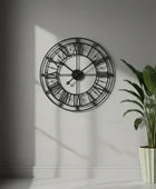 Large wall clock with Roman numeral design on a gray wall next to a potted plant.