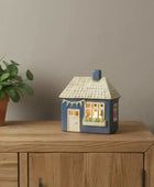 Decorative house-shaped lamp on a wooden surface with a plant beside it.