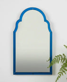 Arched mirror with a blue frame on a white wall with a plant to the right.