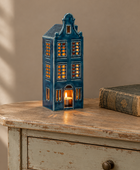 A blue ceramic Dutch house-shaped candle holder with warm glowing lights inside, displayed on a distressed wooden table next to an old book against a beige wall.