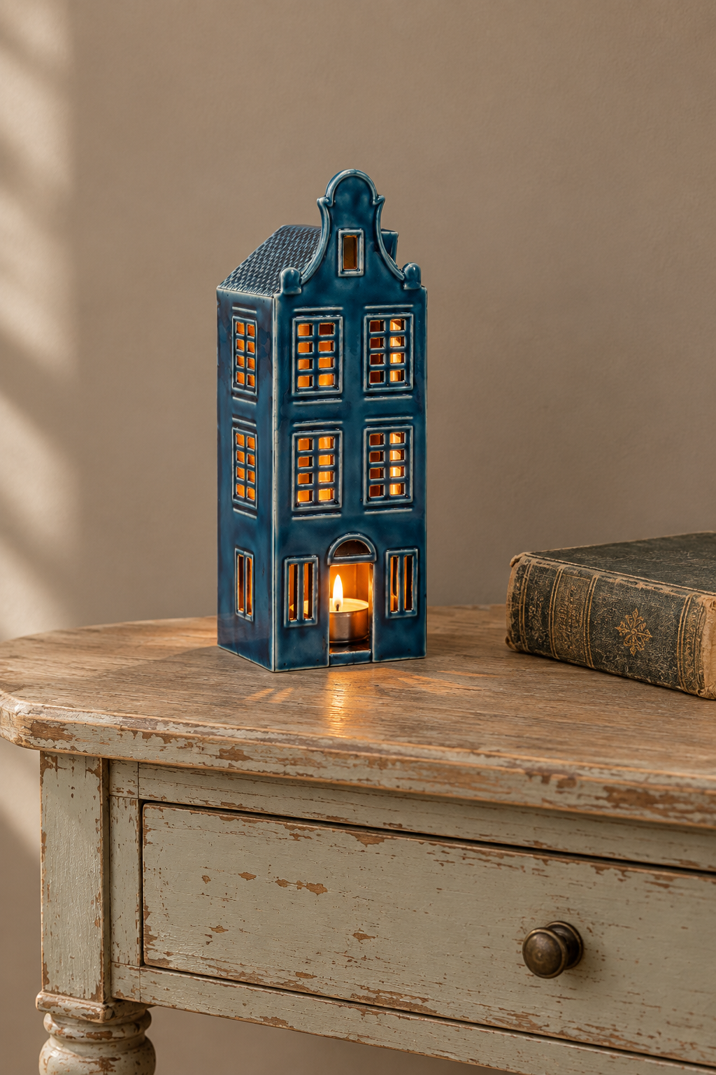 A blue ceramic Dutch house-shaped candle holder with warm glowing lights inside, displayed on a distressed wooden table next to an old book against a beige wall.