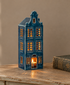 A miniature Dutch house-shaped tealight candle holder with glowing warm lights inside, displayed on a rustic wooden table next to an antique book against a beige wall.