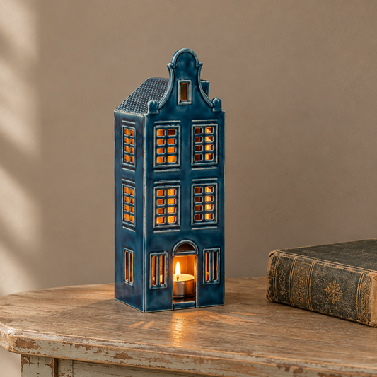 A miniature Dutch house-shaped tealight candle holder with glowing warm lights inside, displayed on a rustic wooden table next to an antique book against a beige wall.