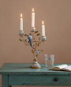 A decorative three-arm candelabra with a gold-toned branch and bird motif, decorated with blue and white porcelain-style accents, holding three lit white taper candles on a rustic wooden table. An open book and a glass of water sit beside it against a warm beige wall.