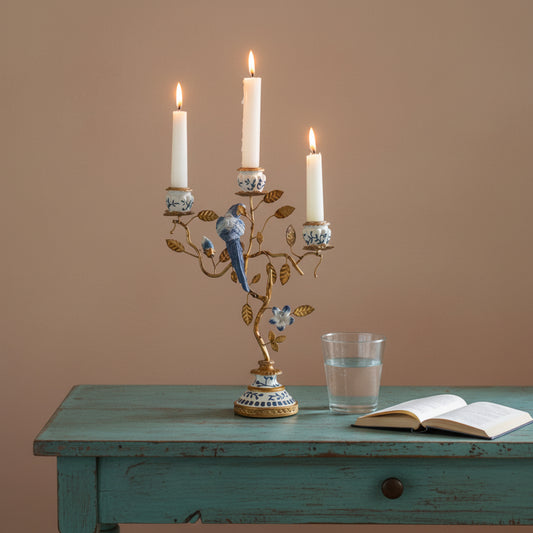A decorative three-arm candelabra with a gold-toned branch and bird motif, decorated with blue and white porcelain-style accents, holding three lit white taper candles on a rustic wooden table. An open book and a glass of water sit beside it against a warm beige wall.