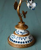 An ornate vintage-style metal and ceramic flower holder or candle holder, featuring a gold-finished branch with a white and blue flower, set on a decorative white and blue patterned base against a rustic wooden background.