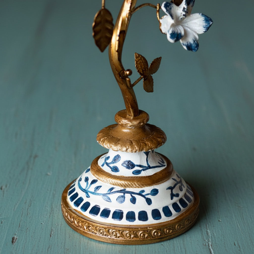An ornate vintage-style metal and ceramic flower holder or candle holder, featuring a gold-finished branch with a white and blue flower, set on a decorative white and blue patterned base against a rustic wooden background.