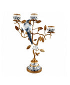 An ornate three-light candelabra designed as a bird perched on a flowering branch, featuring a gold-finished metal frame with blue and white porcelain-style details on the bird, flowers, and base, set against a plain white background.