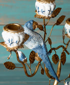 A decorative metal candelabra featuring a blue bird perched on a gold-toned branch structure, with white candle holders decorated with blue floral motifs and gold trim, set against a turquoise wooden background.