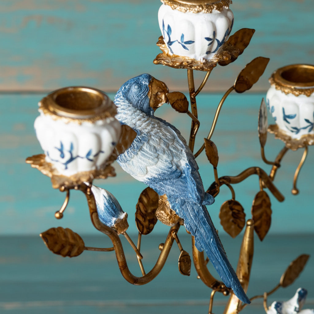 A decorative metal candelabra featuring a blue bird perched on a gold-toned branch structure, with white candle holders decorated with blue floral motifs and gold trim, set against a turquoise wooden background.