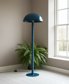 A tall floor lamp with a dome-shaped shade and a ribbed, spiral-like stem in a deep teal blue, standing on a white wood floor next to a potted green fern in a room with beige walls and a window.