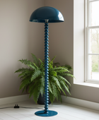 A tall floor lamp with a dome-shaped shade and a vertically ribbed stem, painted in a deep teal blue, standing in a bright room next to a window with a green fern plant in the background.