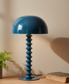Blue table lamp with a textured base on a wooden surface next to a plant and book against a beige wall.