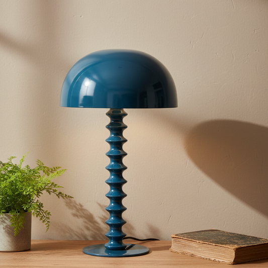 Blue table lamp with a textured base on a wooden surface next to a plant and book against a beige wall.