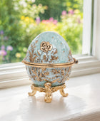 An ornate light blue and gold decorative egg-shaped box with floral detailing, displayed on a white windowsill with a blurred garden background.