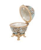 An ornate open decorative egg-shaped box with a light blue enamel exterior, gold-tone metal detailing, and a small gold pedestal base, shown on a plain white background.
