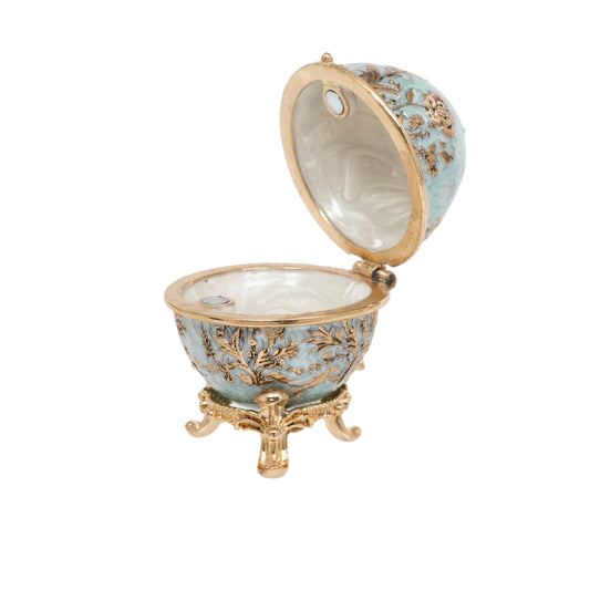 An ornate open decorative egg-shaped box with a light blue enamel exterior, gold-tone metal detailing, and a small gold pedestal base, shown on a plain white background.