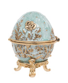 A decorative egg-shaped box with a light aqua enamel finish and ornate gold-tone floral engravings, resting on a matching gold-tone stand against a plain white background.