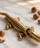 A polished gold-toned metal alligator figurine resting on a white marble surface, surrounded by scattered hazelnuts.