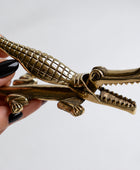 A brass crocodile-shaped door stopper held by a hand against a plain light gray background.