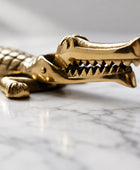 A metallic gold-toned crocodile-shaped accessory, shown in close-up on a neutral gray surface with its mouth open, revealing sharp teeth.