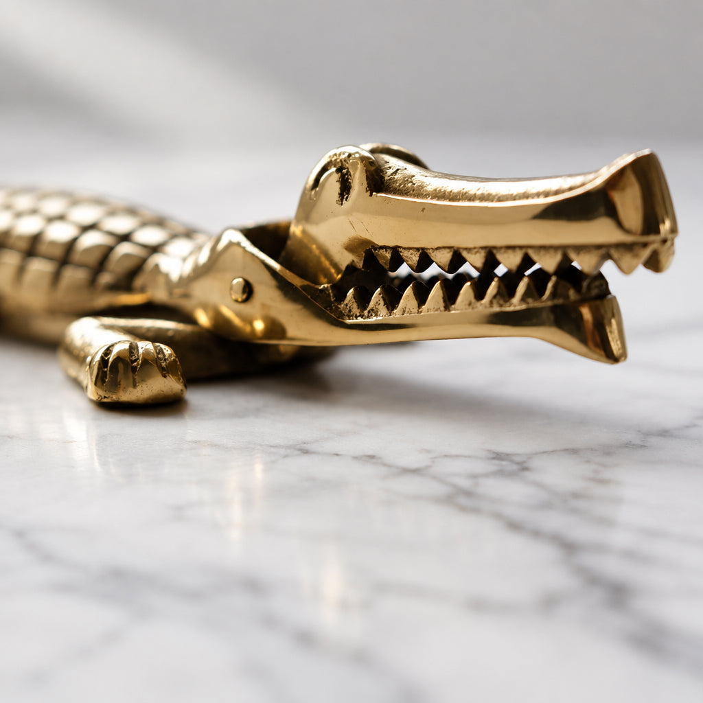 A metallic gold-toned crocodile-shaped accessory, shown in close-up on a neutral gray surface with its mouth open, revealing sharp teeth.