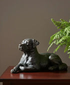 Statue of a lying dog on a wooden surface with a plant in the background