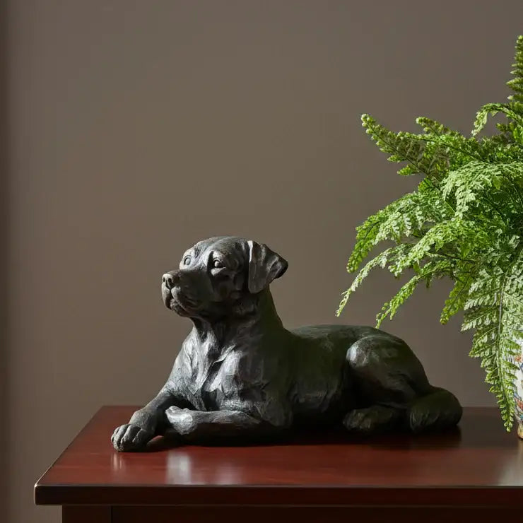 Statue of a lying dog on a wooden surface with a plant in the background