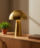 Gold table lamp on a wooden surface with a plant and book in a room with a window.