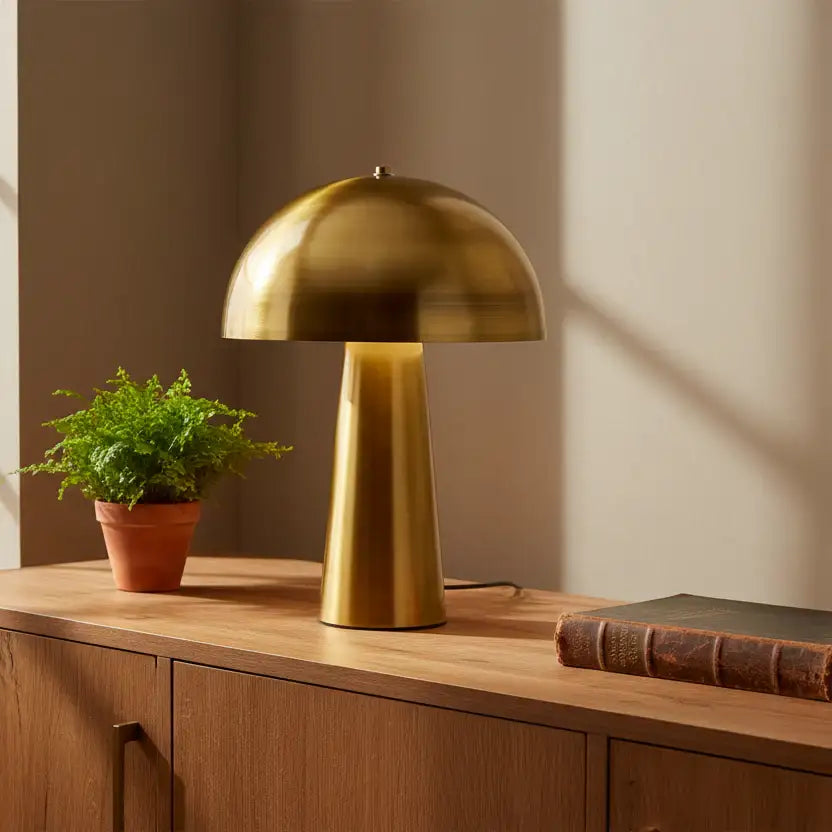 Gold table lamp on a wooden surface with a plant and book in a warm setting